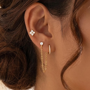 Puede incluir: Un conjunto de joyería para la oreja en tono dorado que incluye un pendiente de flor de cuatro pétalos con piedras claras en la parte superior y un pendiente de piedra única conectado por una fina cadena dorada a un aro pequeño con piedras.