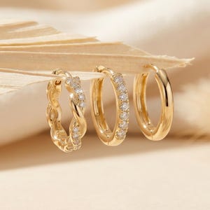 May include: Three gold hoop earrings. One has a twisted design with small clear stones, another has a row of clear stones, and the third is a plain gold hoop. The earrings are displayed on a neutral background.