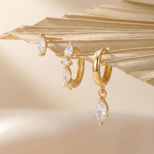 May include: A selection of gold-tone earrings featuring clear gemstones. The earrings include a stud, a small hoop with multiple stones, and a larger hoop with a dangling teardrop-shaped stone. The earrings are presented on a dried palm leaf.