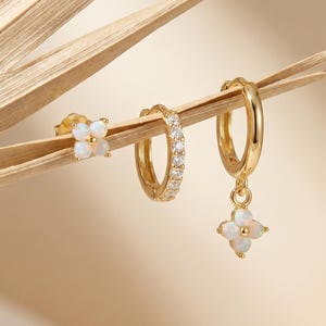 Puede incluir: Un conjunto de pendientes dorados. El conjunto incluye un pendiente de botón con un diseño de flor de cuatro pétalos, un pendiente de aro pequeño con piedras transparentes y un pendiente de aro más grande con un dije de flor de cuatro pétalos colgante. Los pendientes se exhiben sobre una hoja de palma seca.