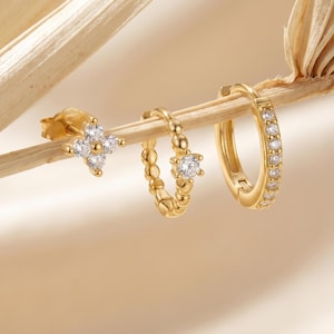 Puede incluir: Tres pendientes dorados con piedras preciosas transparentes. Uno es un pendiente de botón con un diseño de flor de cuatro pétalos. El segundo es un aro pequeño con una sola piedra preciosa. El tercero es un aro más grande con múltiples piedras preciosas.