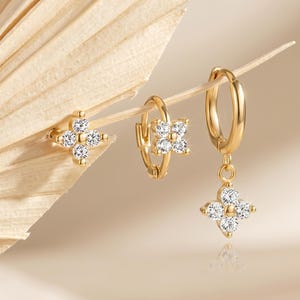 Puede incluir: Un conjunto de tres pendientes dorados con un diseño de flor de cuatro pétalos. Un pendiente es un botón, otro es un aro pequeño con un dije de flor fijo y el tercero es un aro pequeño con un dije de flor colgante, todos con piedras blancas.