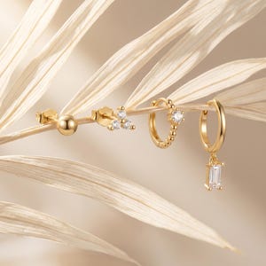 May include: A set of four gold-toned earrings consisting of a small ball stud, a three-stone cluster stud, a beaded hoop earring with a single clear stone, and a smooth hoop earring featuring a dangling rectangular clear baguette-cut stone.