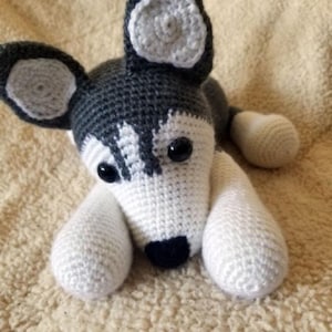 May include: A handmade crochet husky dog toy. The toy features a white and gray color scheme, with black eyes and nose. The dog is lying down, with its paws extended. The ears have a unique spiral design.