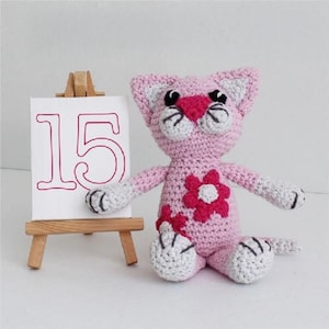 May include: A handmade pink crochet cat with white and gray accents, featuring a heart-shaped nose and flower embellishments. The cat sits next to a small wooden easel holding a canvas with the number 15.
