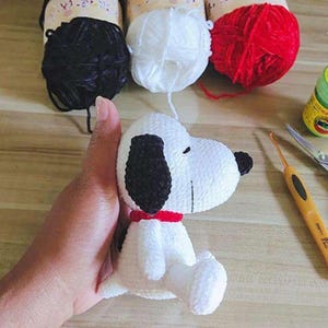 May include: A crocheted Snoopy doll in white and black, with a red collar, is held in a hand. Balls of black, white, and red yarn are visible, alongside a crochet hook and scissors. The doll is a well-known cartoon character.