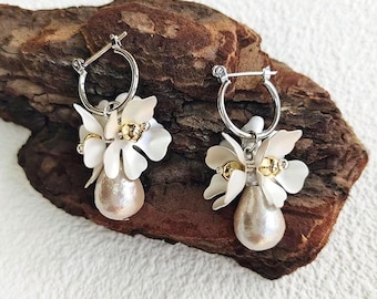 May include: A pair of silver hoop earrings with white flower and pearl drop accents. Each earring features a cluster of white flower petals, a small gold bead, and a large, pearlescent drop. The earrings are displayed on a piece of brown bark.