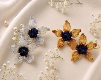 May include: Two silver-toned and two gold-toned flower-shaped brooches, each featuring five delicate wire-wrapped petals and a dark blue beaded center, arranged alongside small white baby's breath flowers and a single pearl.