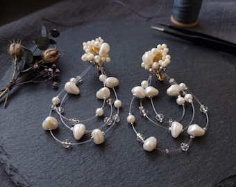 May include: A pair of drop earrings featuring clusters of small white beads at the top, with multiple clear nylon threads hanging down. Each thread is adorned with white teardrop-shaped beads and small clear faceted crystals in a tiered design.