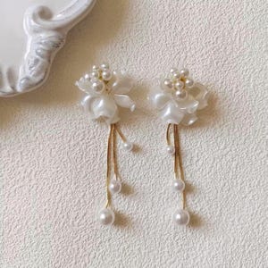 May include: A pair of white floral earrings with pearl accents. Each earring features a cluster of small pearls at the center of the flower and dangling strands of pearls. The earrings have gold-colored chains and are set against a textured white background.