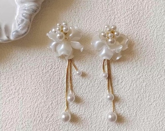May include: A pair of white floral earrings with pearl accents. Each earring features a cluster of small pearls at the center of the flower and dangling strands of pearls. The earrings have gold-colored chains and are set against a textured white background.