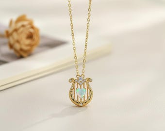 May include: A gold-colored necklace with a harp-shaped pendant. The pendant features a heart-shaped opal stone and is adorned with small, clear gemstones. The necklace is displayed on a white surface.