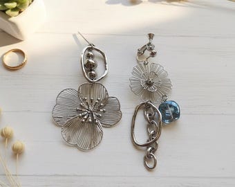 May include: Two silver-toned earrings. One features a large, wire-frame flower and a smaller oval with three beads. The other has a wire-frame flower, a blue glass bead, and a chain. The earrings are on a white wooden surface.
