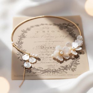 Può includere: Una delicata collana girocollo dorata con dettagli di fiori bianchi e perle. Gli elementi floreali sono raggruppati sul lato destro, con alcuni petali e perle pendenti sul lato sinistro. La collana poggia su uno sfondo di carta vintage.