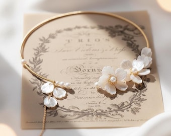 May include: A delicate gold-toned choker necklace featuring white flower and pearl accents. The floral elements are clustered on the right side, with a few dangling petals and pearls on the left. The necklace rests on a vintage paper background.