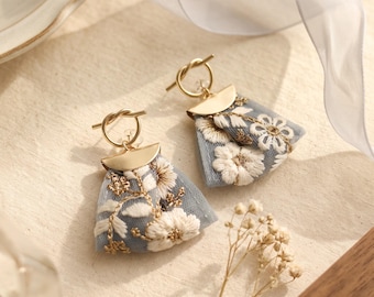 May include: A pair of dangle earrings featuring a gold-toned metal knot and semi-circle top piece attached to a triangular piece of light blue tulle fabric. The tulle is decorated with white floral embroidery and gold-toned thread accents.