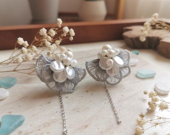 May include: A pair of silver lace earrings with white pearl accents and dangling chains. The earrings have a floral design with multiple pearl sizes and are displayed on a marble surface.