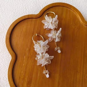 May include: A pair of gold hoop earrings and a matching clip-on earring, each adorned with delicate white flower petals and sparkling crystals. The earrings dangle with a small chain and a pearlescent bead. The earrings are displayed on a wooden tray.
