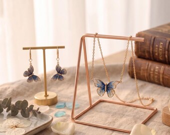 May include: A gold-toned metal T-bar earring stand displaying a pair of butterfly earrings with blue and white enamel, and a matching gold-toned triangular necklace stand holding a delicate chain necklace with a blue and white butterfly pendant.