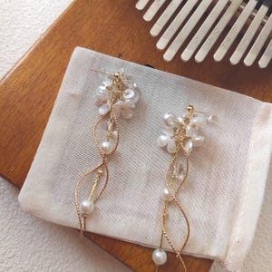 May include: A pair of elegant earrings featuring white flower accents, clear beads, and dangling pearls. The earrings have a gold-colored wire design and are displayed on a white cloth pouch. A wooden surface and a kalimba are in the background.