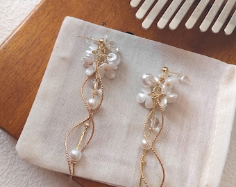 May include: A pair of elegant earrings featuring white flower accents, clear beads, and dangling pearls. The earrings have a gold-colored wire design and are displayed on a white cloth pouch. A wooden surface and a kalimba are in the background.