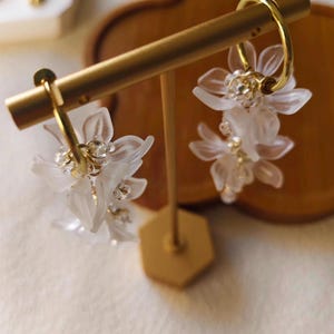 May include: A pair of gold hoop earrings with delicate, white, flower-shaped pendants. The earrings are displayed on a gold stand, showcasing their intricate design and sparkling accents. The earrings are a floral-themed accessory.