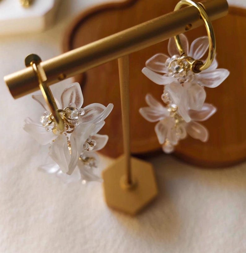 May include: A pair of gold hoop earrings with delicate, white, flower-shaped pendants. The earrings are displayed on a gold stand, showcasing their intricate design and sparkling accents. The earrings are a floral-themed accessory.