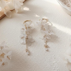 May include: A pair of elegant white floral earrings with gold-colored hoops and delicate chain accents. The earrings feature cascading white flower petals and small, sparkling embellishments, creating a graceful and feminine design. Perfect for special occasions.