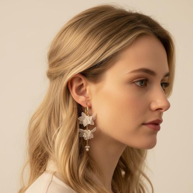 May include: Gold hoop earrings with three white fabric flower pendants and a small pearl at the bottom. The earrings are worn by a person with blonde hair.