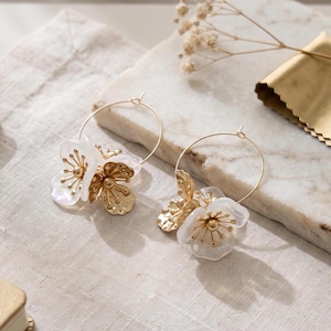 May include: A pair of gold hoop earrings featuring white translucent flower petals and gold-toned stamens. Each earring is designed with a thin circular wire hoop that supports the floral arrangement, combining metallic gold elements with white accents.
