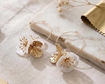 May include: A pair of gold hoop earrings featuring white translucent flower petals and gold-toned stamens. Each earring is designed with a thin circular wire hoop that supports the floral arrangement, combining metallic gold elements with white accents.
