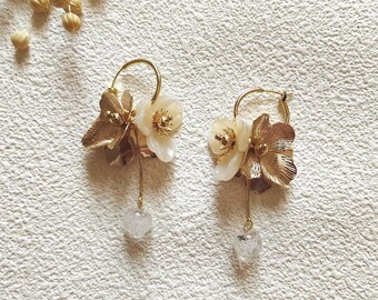 May include: A pair of gold-toned hoop earrings with floral and leaf accents. Each earring features a cluster of cream-colored flowers, gold leaves, and a dangling clear, heart-shaped charm. The earrings are set against a textured white background.