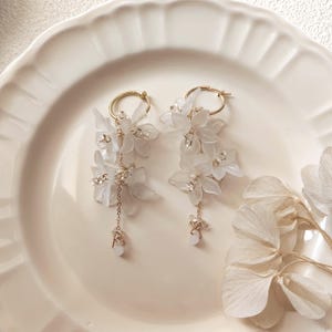 May include: A pair of elegant white floral earrings with gold-colored hoops. Each earring features delicate white flower clusters, accented with small crystals and dangling gold chains. The earrings are displayed on a white plate.