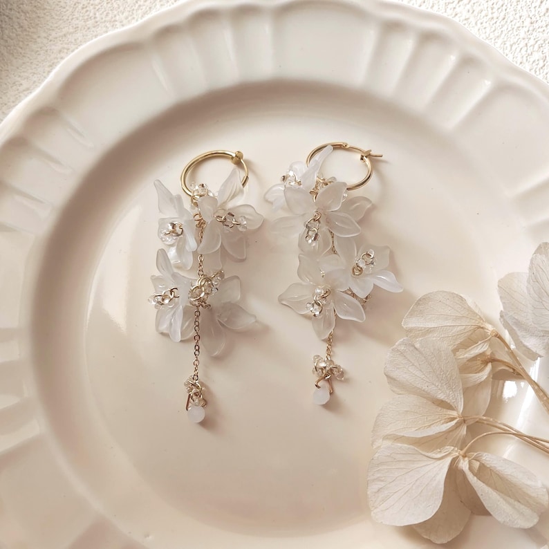 May include: A pair of elegant white floral earrings with gold-colored hoops. Each earring features delicate white flower clusters, accented with small crystals and dangling gold chains. The earrings are displayed on a white plate.