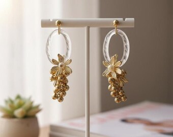 May include: Clear oval earrings with a twisted design, featuring gold-colored floral and grape cluster charms. The earrings are displayed on a white stand, showcasing their intricate details and elegant design.