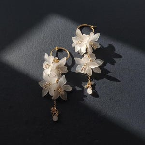 May include: A pair of elegant earrings featuring delicate white flower pendants suspended from gold-colored hoops. The earrings have multiple flower clusters and small dangling accents, set against a dark background.