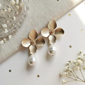 May include: A pair of gold flower earrings with a pearl drop. The earrings feature a four-petal design with a textured finish. The pearls are large and teardrop-shaped. The earrings are displayed on a white surface with floral accents.