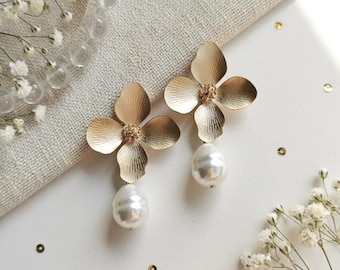 May include: A pair of gold-colored flower earrings with a pearl drop. The earrings have a four-petal design with a textured finish. The pearls are large and teardrop-shaped. The earrings are displayed on a white surface with floral accents.
