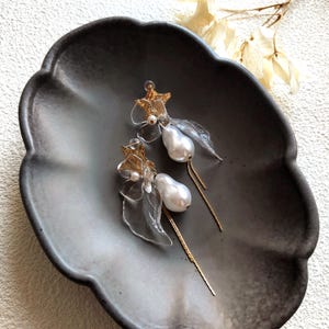 Pode incluir: Um par de brincos pendurados elegantes com grandes pérolas brancas peroladas. Os brincos têm detalhes dourados em forma de estrela e elementos transparentes em forma de folha. Delicadas correntes douradas pendem das pérolas. Os brincos são exibidos em um prato cinza escuro.