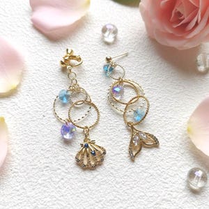 May include: A pair of gold-toned earrings with a mermaid theme. Each earring features multiple gold rings, blue and iridescent crystal beads, and a charm. One earring has a seashell charm, the other a mermaid tail.