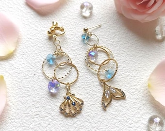 May include: A pair of gold-toned earrings with a mermaid theme. Each earring features multiple gold rings, blue and iridescent crystal beads, and a charm. One earring has a seashell charm, the other a mermaid tail.
