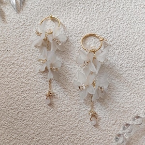 May include: A pair of elegant white floral earrings with gold-colored hoops and delicate chain accents. The earrings feature multiple tiers of white flower petals and small, sparkling embellishments, creating a sophisticated and feminine design.