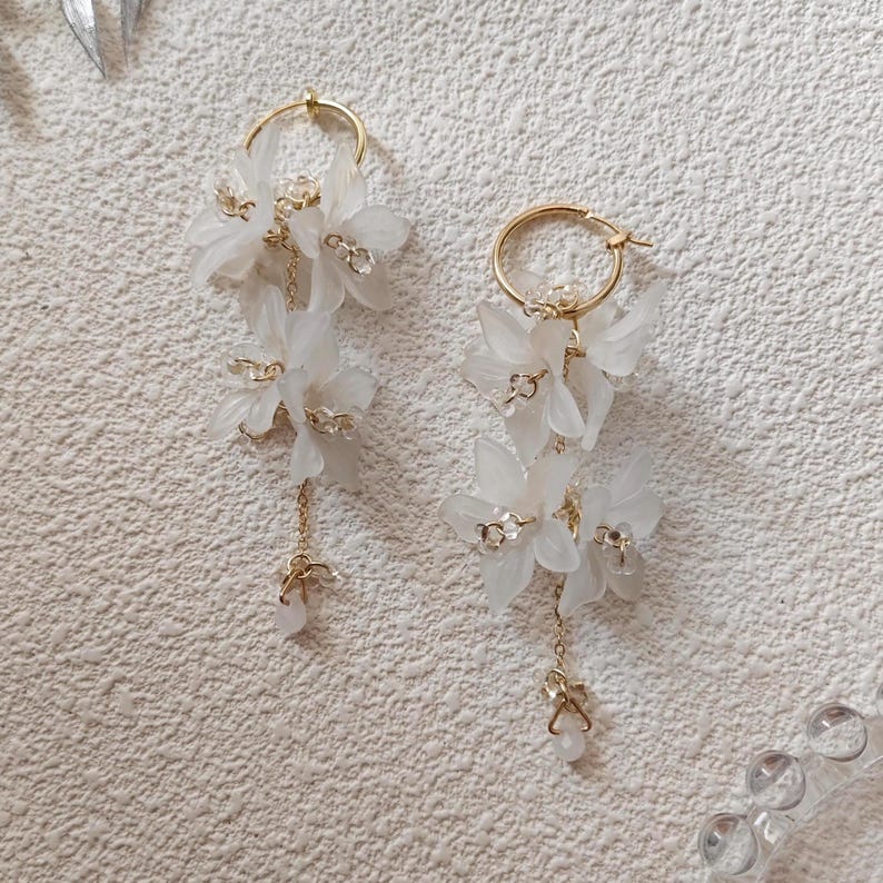 May include: A pair of elegant white floral earrings with gold-colored hoops and delicate chain accents. The earrings feature multiple tiers of white flower petals and small, sparkling embellishments, creating a sophisticated and feminine design.