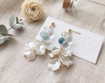 May include: A pair of dangle earrings featuring gold-tone posts, clusters of small translucent blue and clear beads, and multiple white petal-shaped charms. The earrings are displayed on a white rectangular card with an embossed floral design.