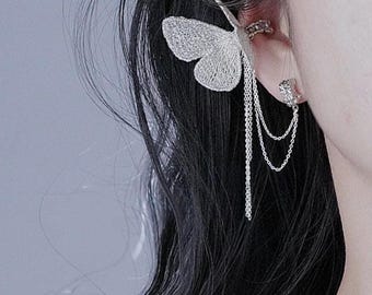 May include: A silver ear cuff featuring a delicate butterfly design. The butterfly has a lacy, openwork pattern and is attached to a series of dangling chains. The cuff wraps around the ear, adding a touch of elegance.