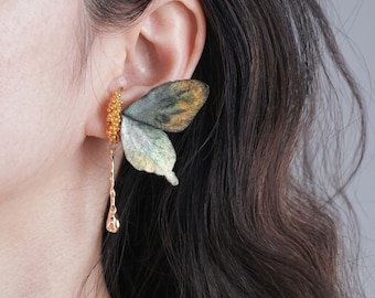 May include: An ear cuff featuring a textured gold-tone band with a small dangling teardrop charm and a large, wing-shaped attachment in shades of muted green, brown, and yellow, designed to sit along the outer edge of the ear cartilage.