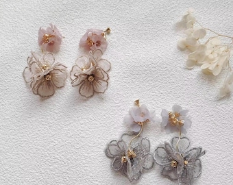 May include: Three pairs of floral earrings. Two pairs feature pink and gold accents, with delicate, layered petals. The third pair has gray petals and gold details. The earrings are set against a white textured background.