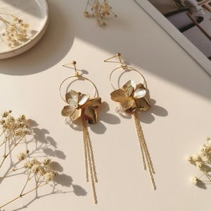 Könnte beinhalten: Ein Paar goldfarbene Blumenohrringe. Jeder Ohrring hat einen Blumenanhänger mit einer herabhängenden Kette und einer klaren Kristallperle. Die Ohrringe sind vor einem neutralen Hintergrund mit getrockneten Blumen platziert.