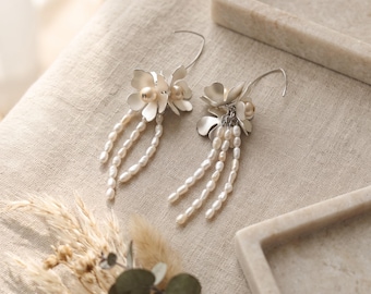 May include: A pair of silver-toned earrings featuring matte metal flower petals, a central round bead, and three dangling strands of small, irregular white freshwater pearls attached to a curved silver-toned ear wire hook for secure placement.