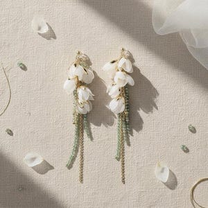 May include: A pair of drop earrings with white fabric flower clusters. The earrings have gold-coloured details and strands of green beads and gold chains, creating a delicate and feminine design.